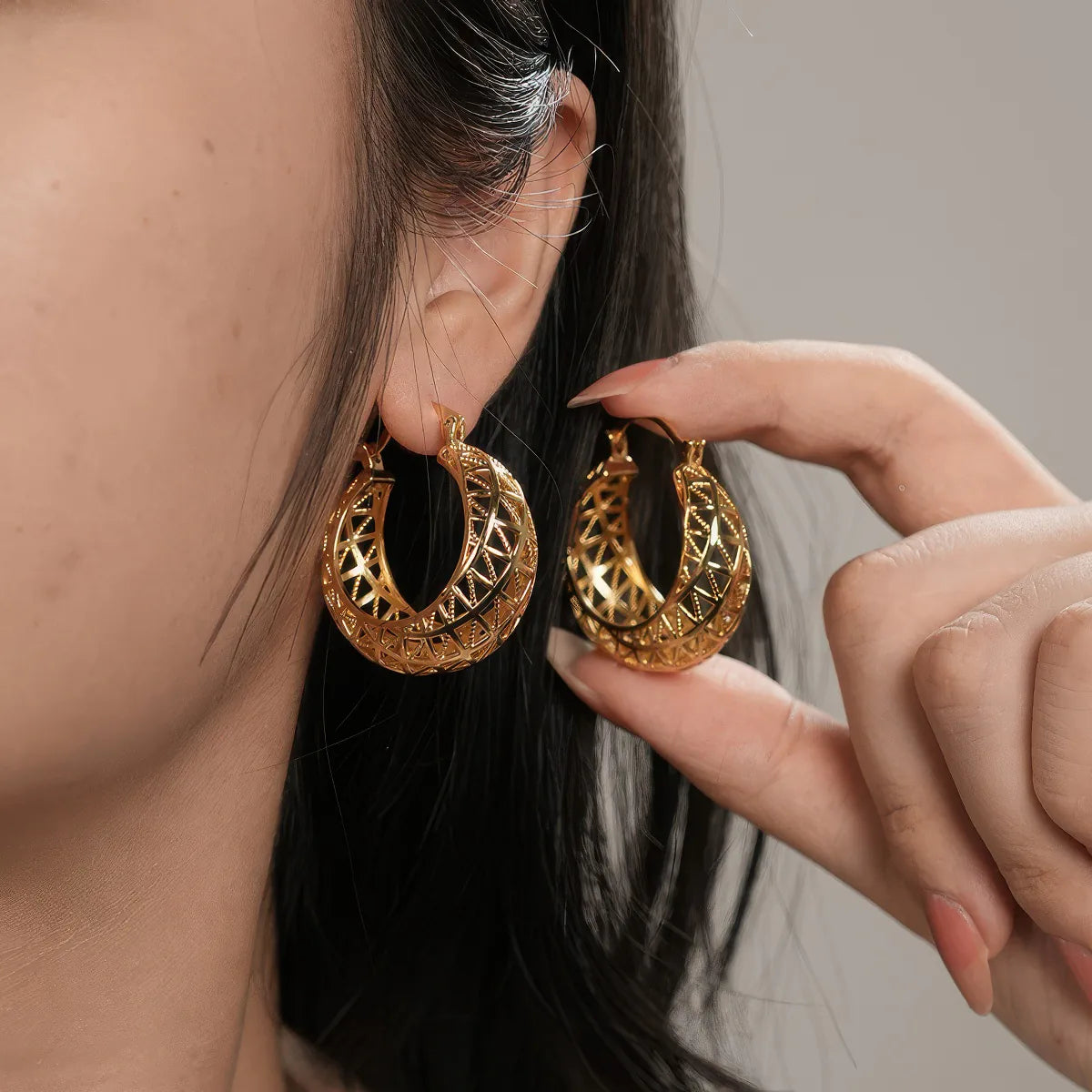 Gold hoop earrings being worn and held by a person against a neutral background. #Style_B