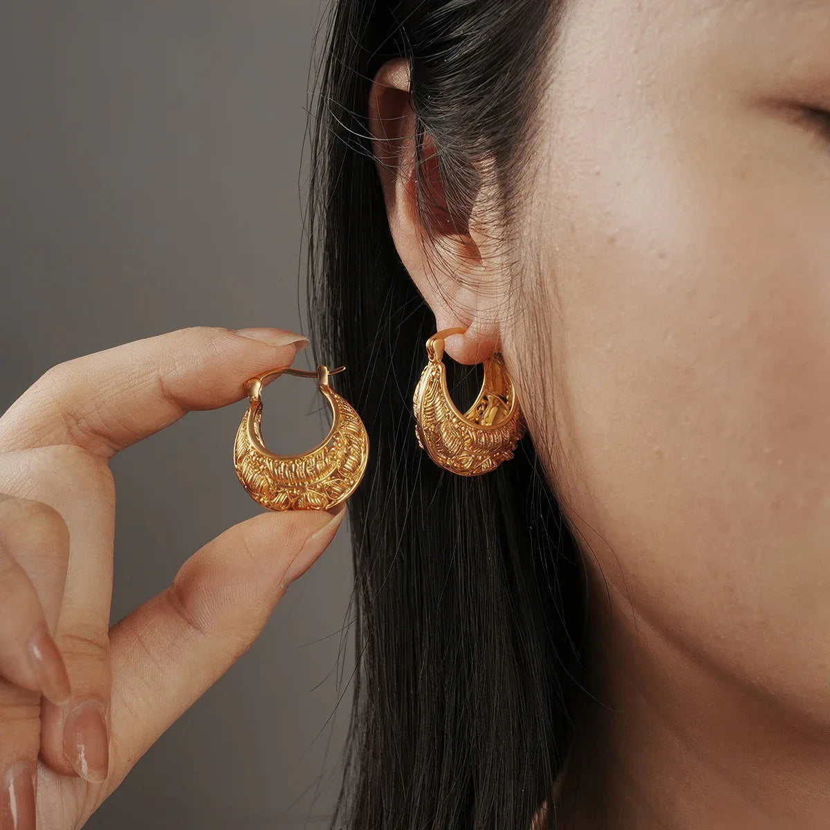 Gold hoop earrings being worn and held by a hand against a neutral background. #Style_C