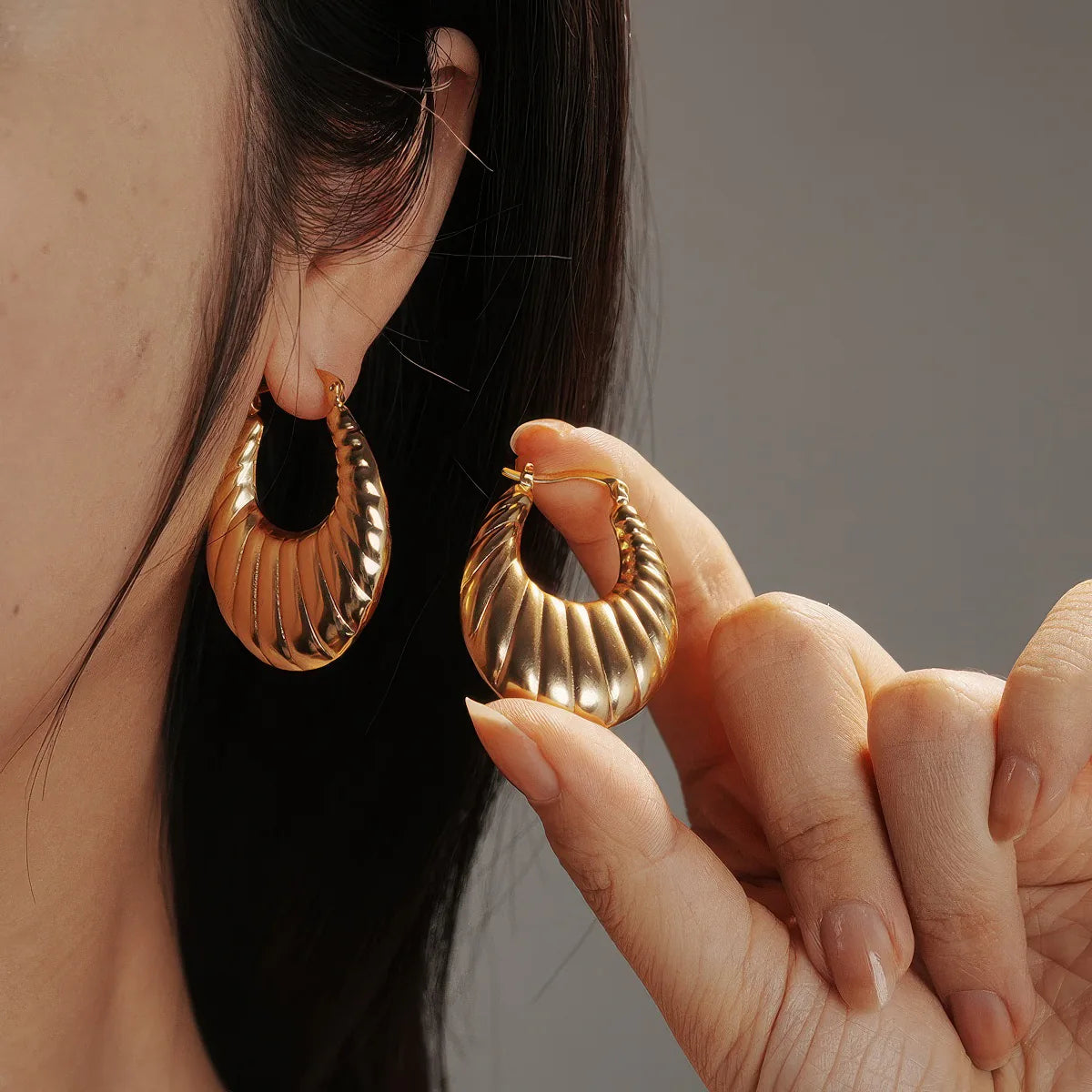 Gold hoop earrings being worn and held by a person against a neutral background. #Style_B