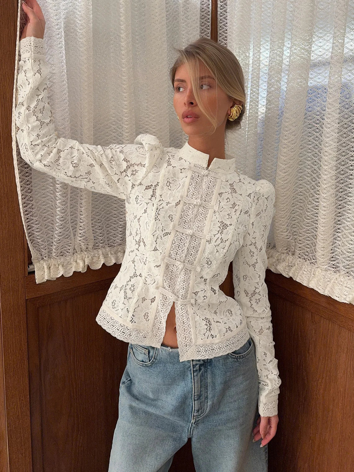 Woman wearing a white lace top and blue jeans standing in front of a textured wall. #Color_White
