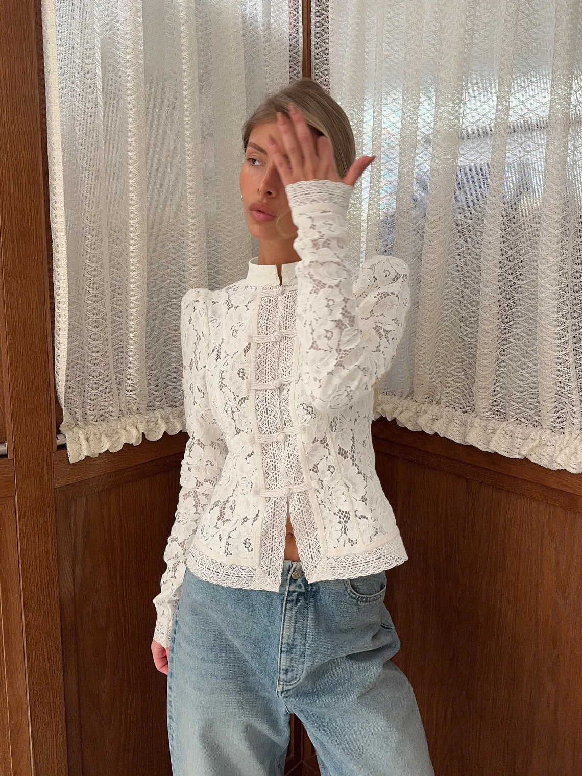 Person wearing a white lace top and blue jeans in front of a window with white curtains. #Color_White