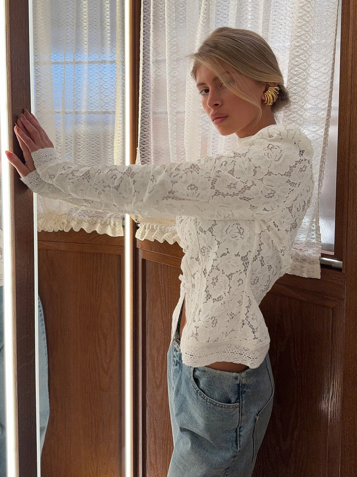 Woman wearing a white lace top and blue jeans standing in front of a window with sheer curtains. #Color_White