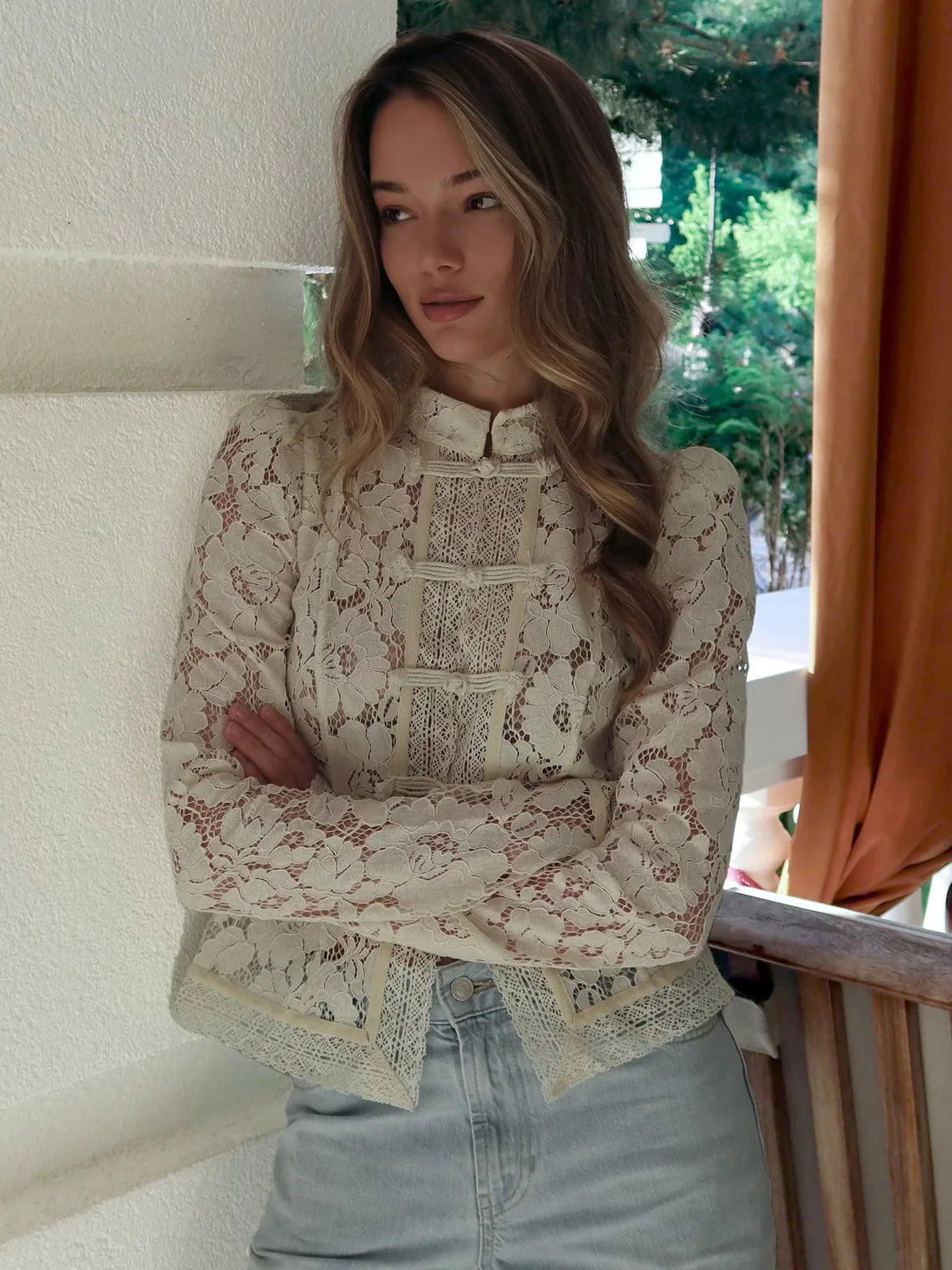 Woman wearing a patterned blouse and light blue jeans standing against a white wall with greenery in the background. #Color_Beige