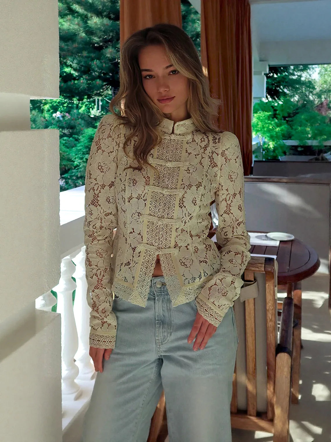 Woman wearing a lace top and light blue jeans indoors with greenery outside. #Color_Beige