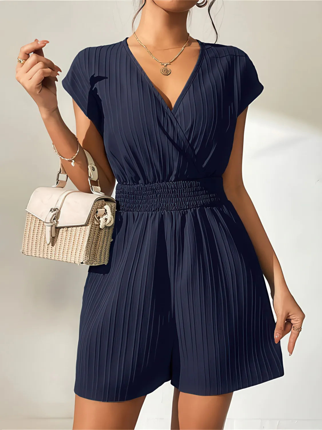 Woman wearing a navy blue romper with a white handbag on a neutral background. #Color_MidnightBlue