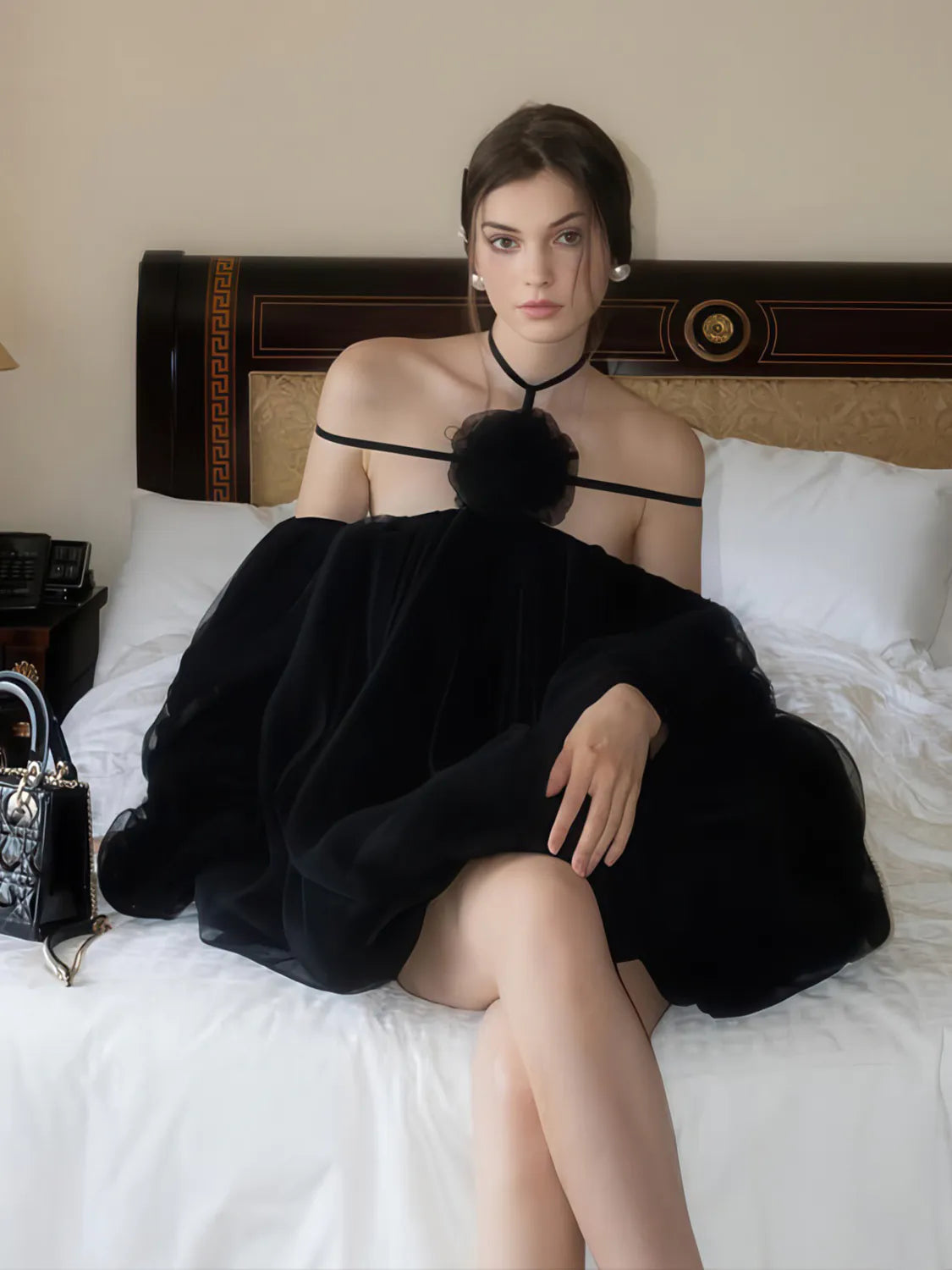 Woman in a black dress sitting on a bed with a handbag beside her. #Color_Black