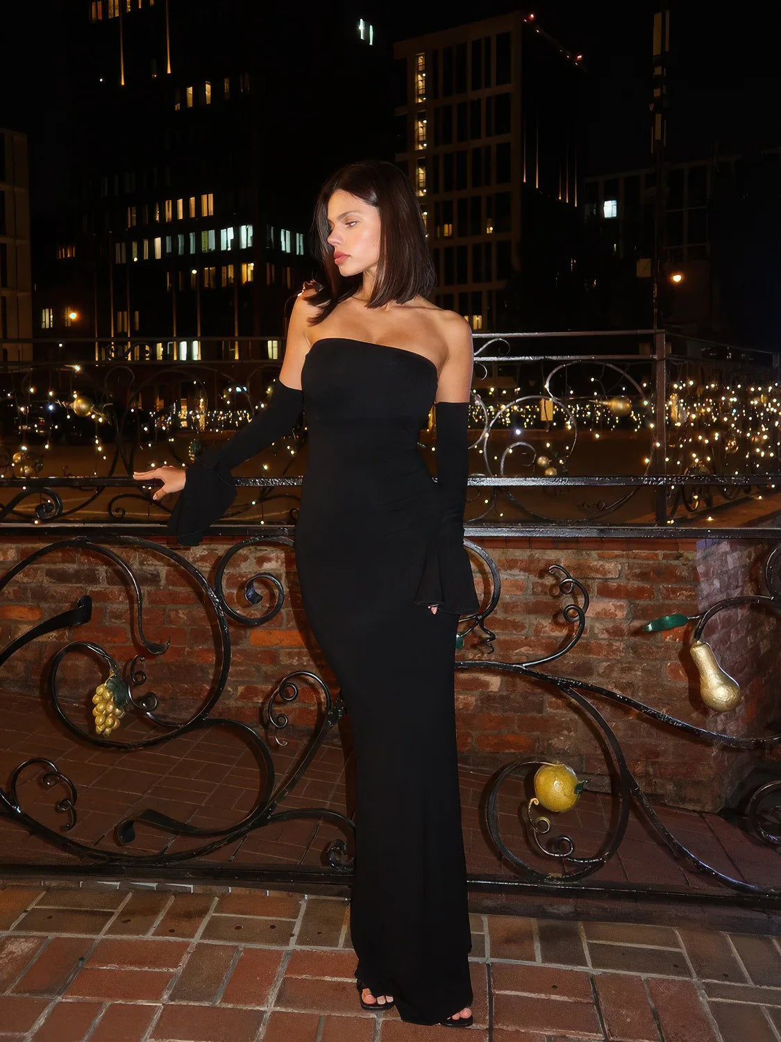 Woman in a black dress standing on a rooftop at night with city lights in the background. #Color_Black