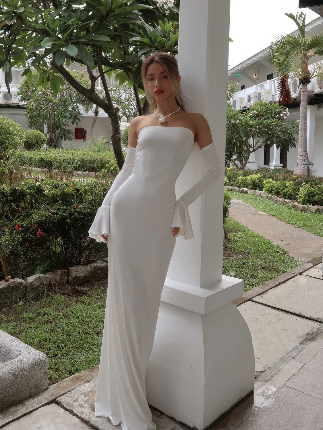 Woman in a white strapless gown standing outdoors near a garden. #Color_White