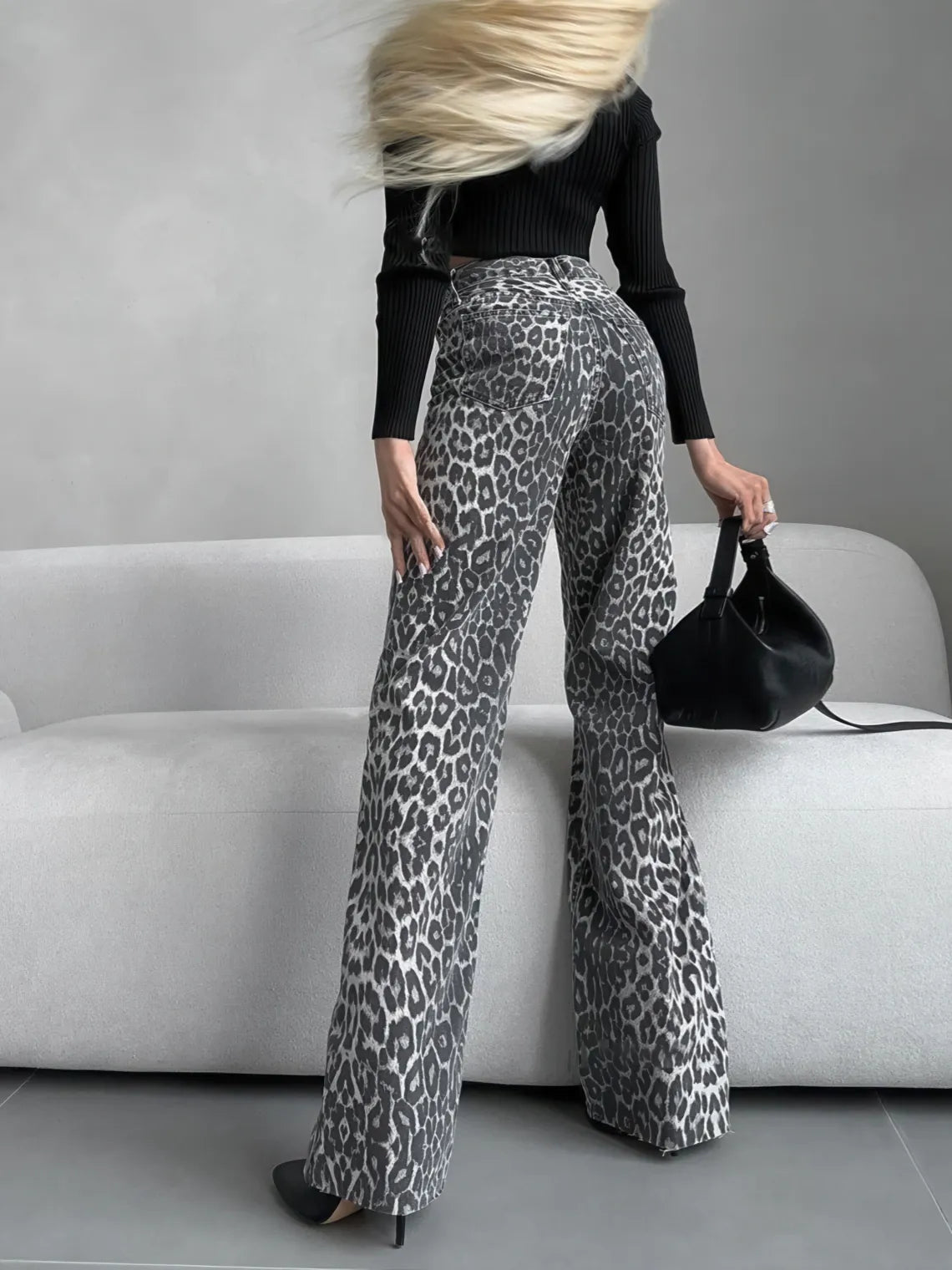Person wearing leopard print pants and a black top, holding a black handbag, sitting on a gray couch. #Color_Gray