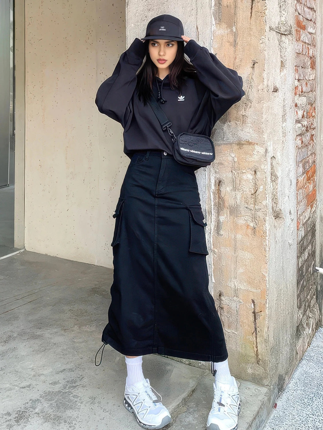 Person wearing a black outfit with a bag and white sneakers, leaning against a wall. #Color_Black