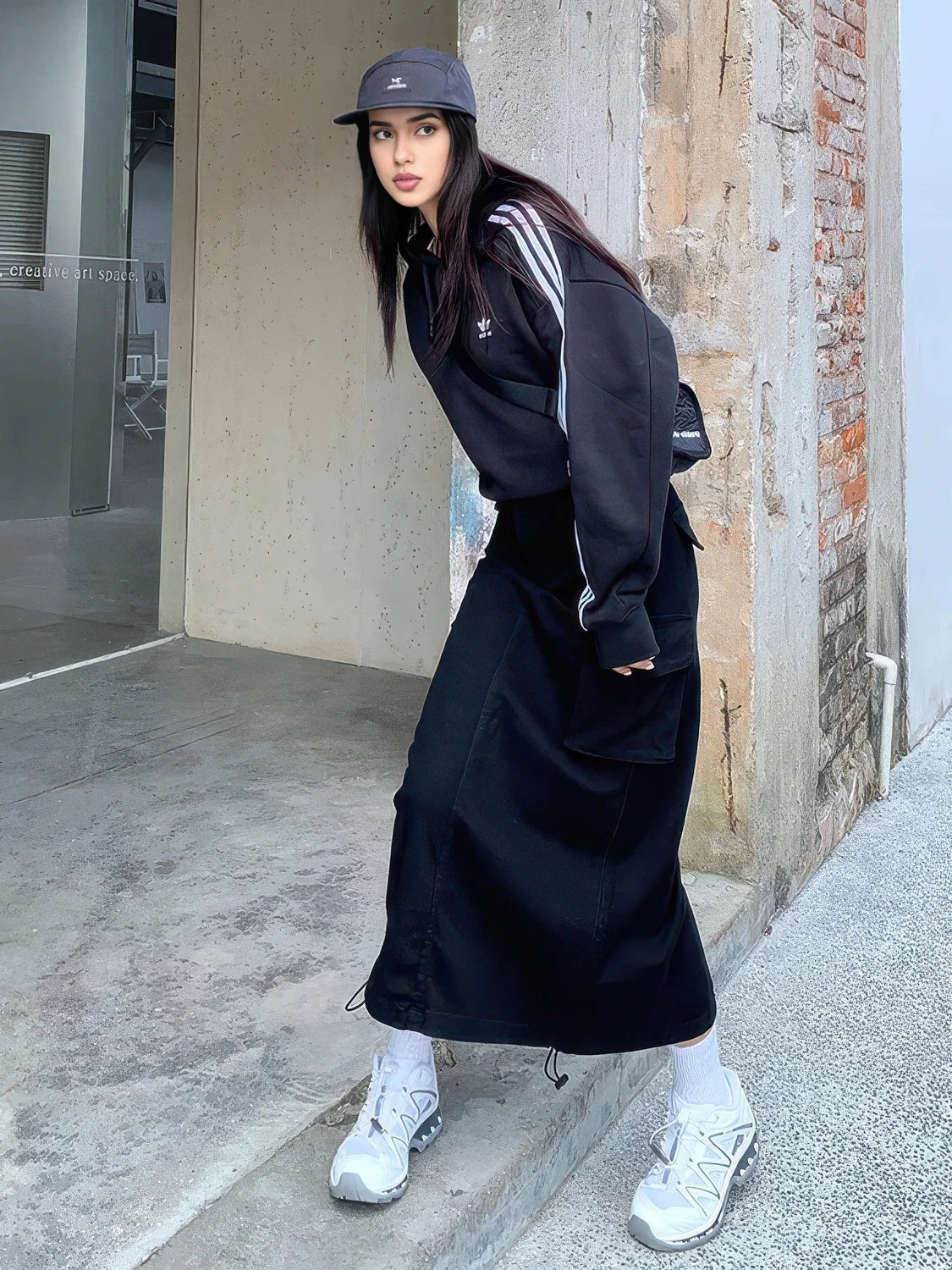 Person wearing a black outfit with white sneakers standing against a wall. #Color_Black
