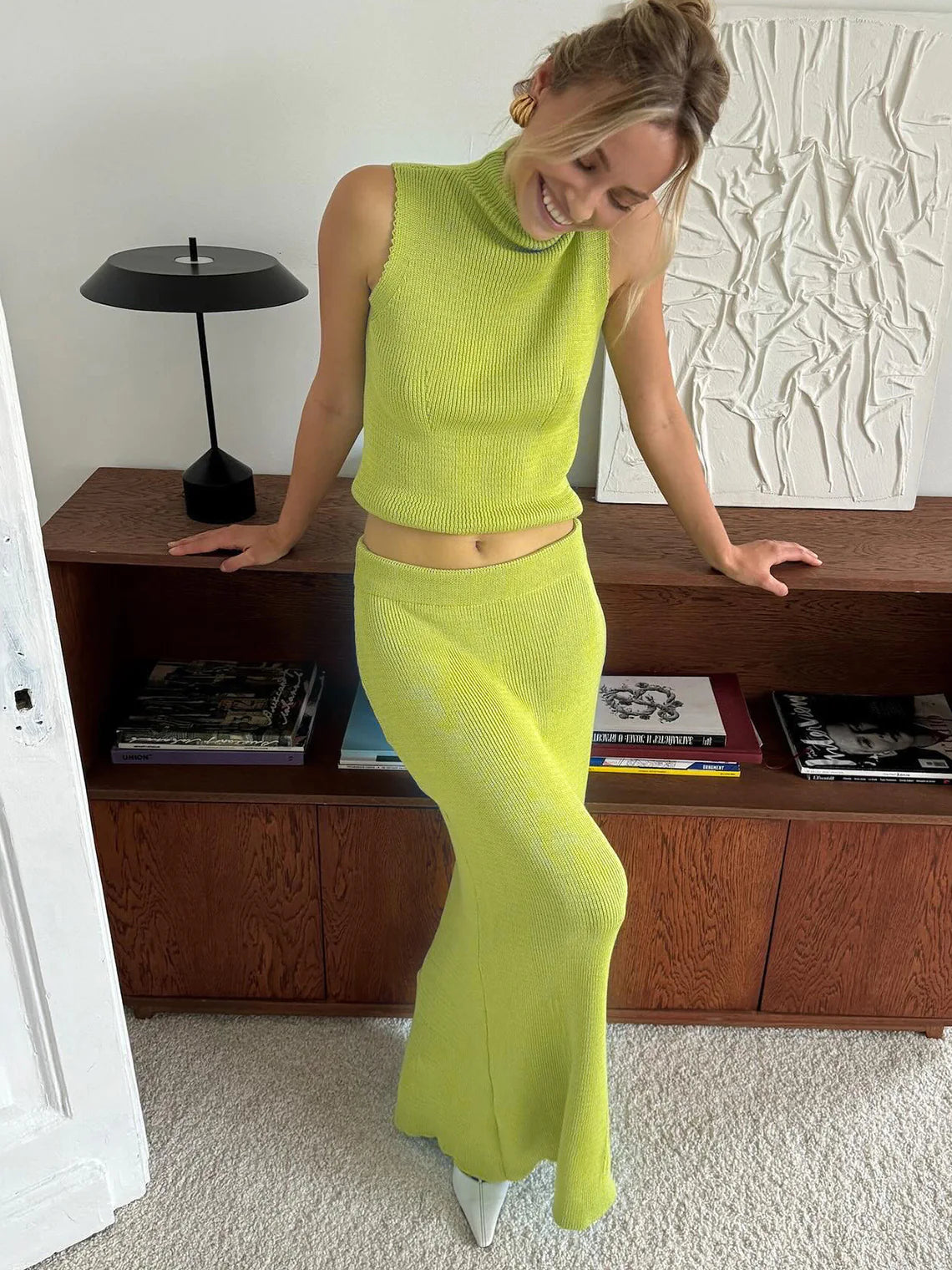 Woman in a bright green outfit standing in a room with a wooden cabinet and lamp. #Color_Green