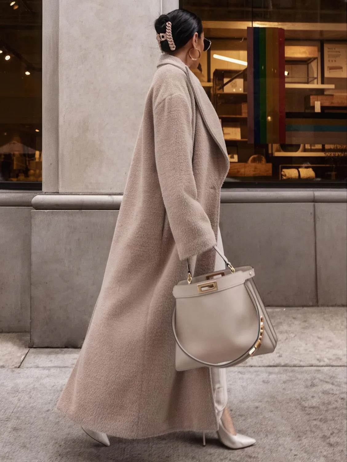 Woman in a long beige coat holding a matching handbag, walking past a store.
