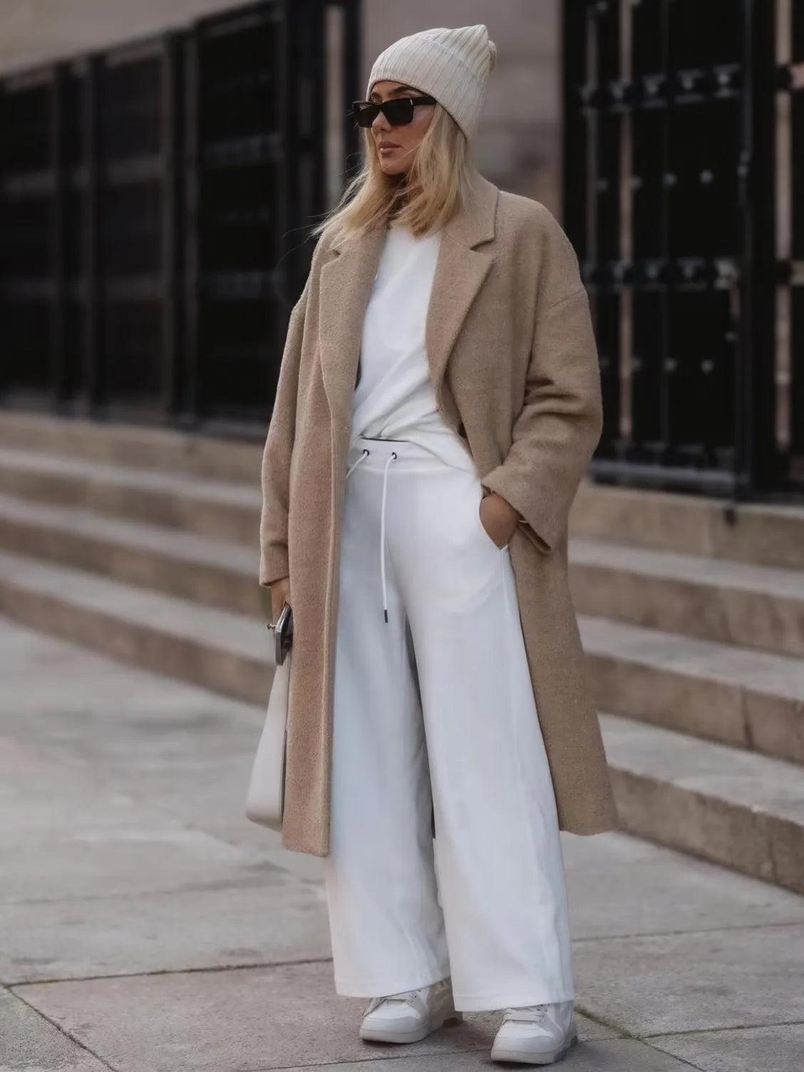 Person wearing a beige coat, white pants, and a beanie outdoors.