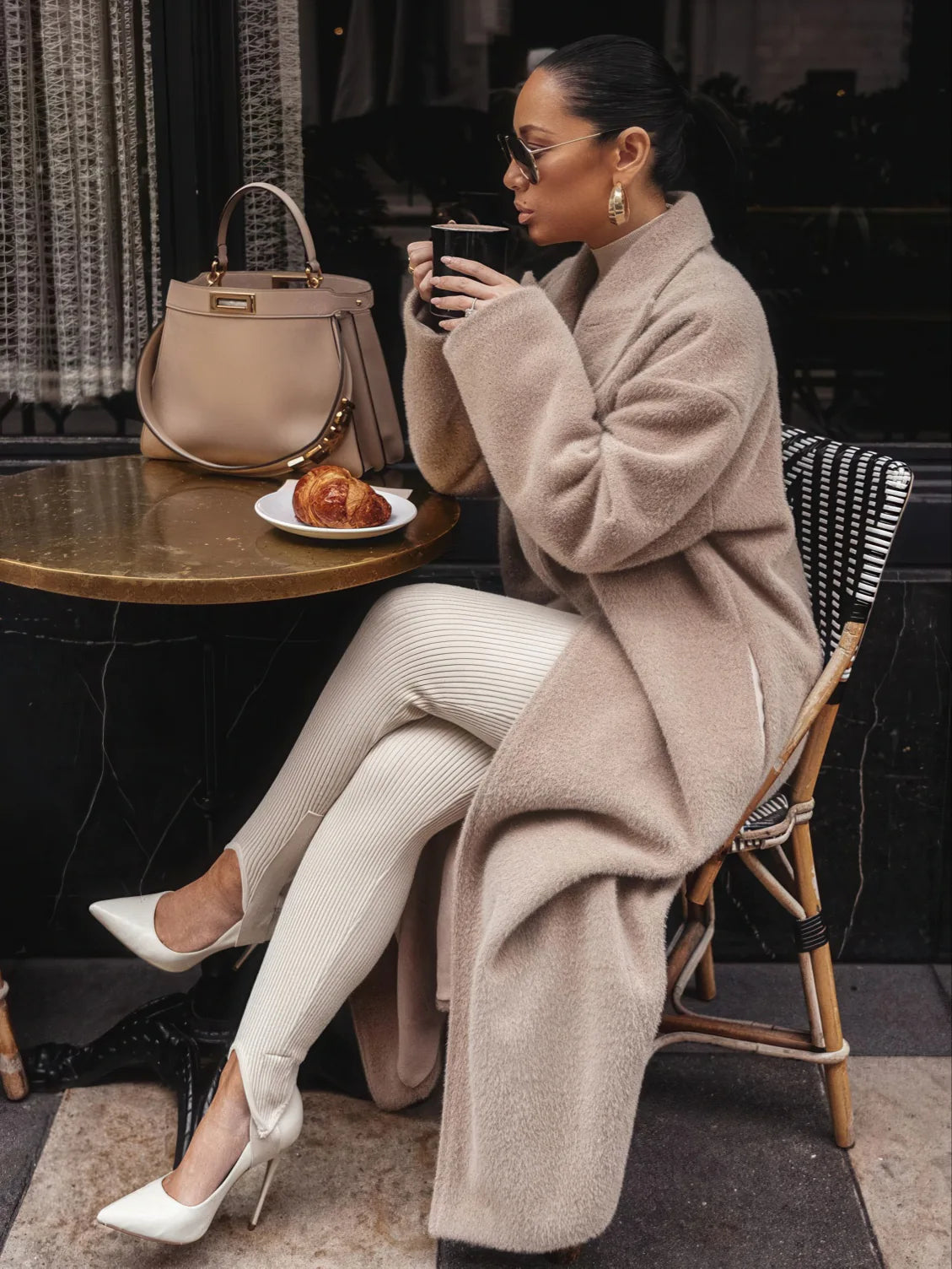 Woman in a beige coat sitting at an outdoor cafe table, holding a cup and plate with a pastry.