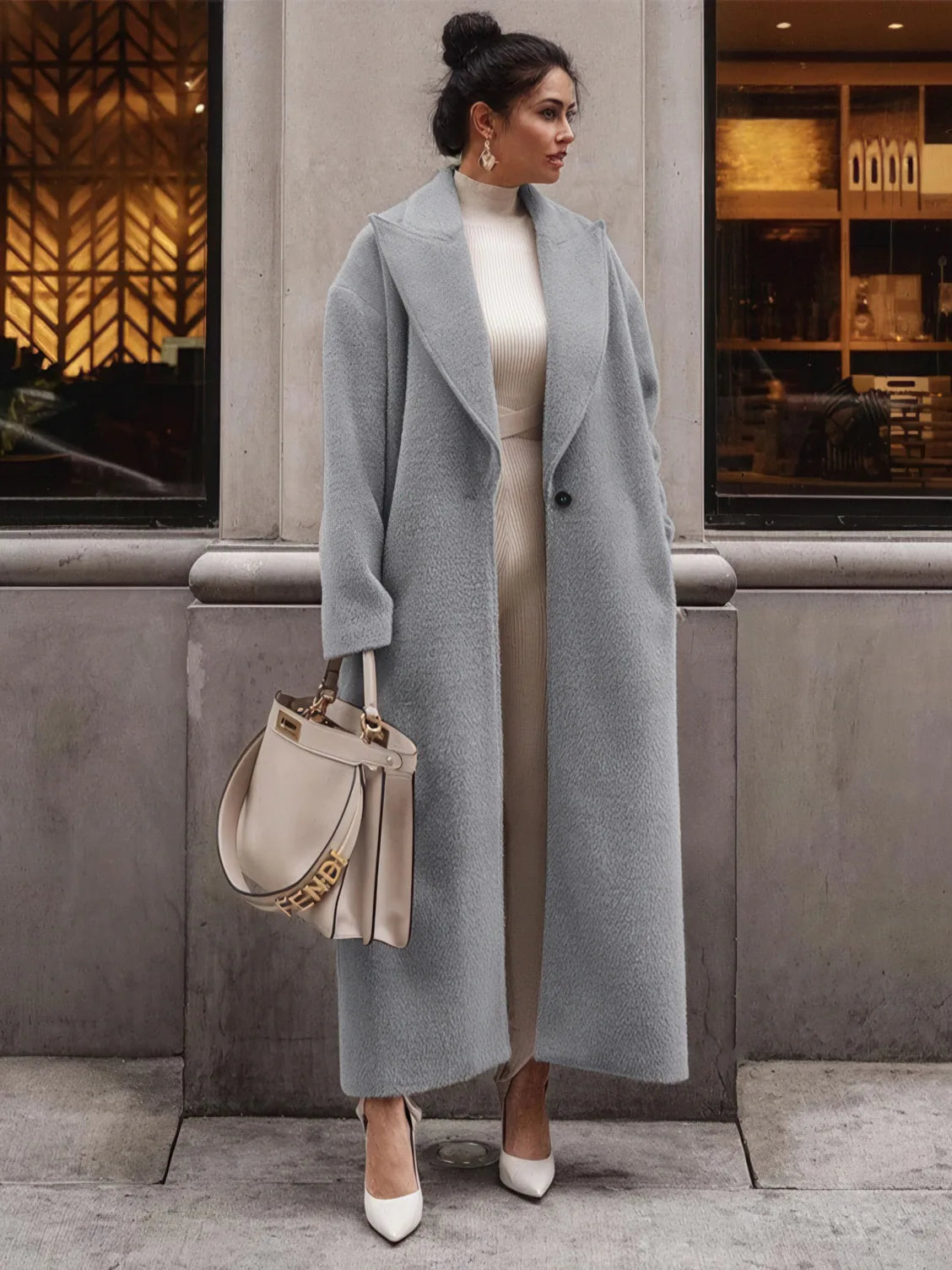 Woman wearing a long gray coat with a neutral handbag, standing in front of a building.