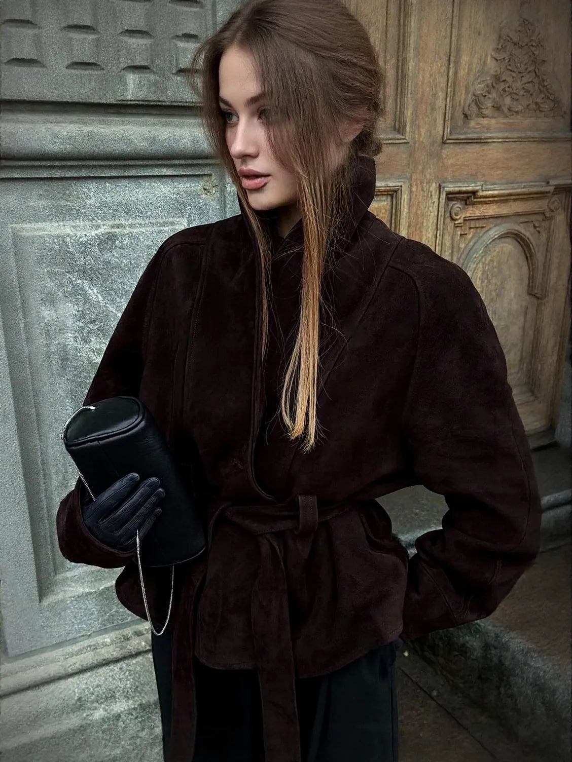 Woman in a brown coat holding a black bag against a textured wall. #Color_Brown