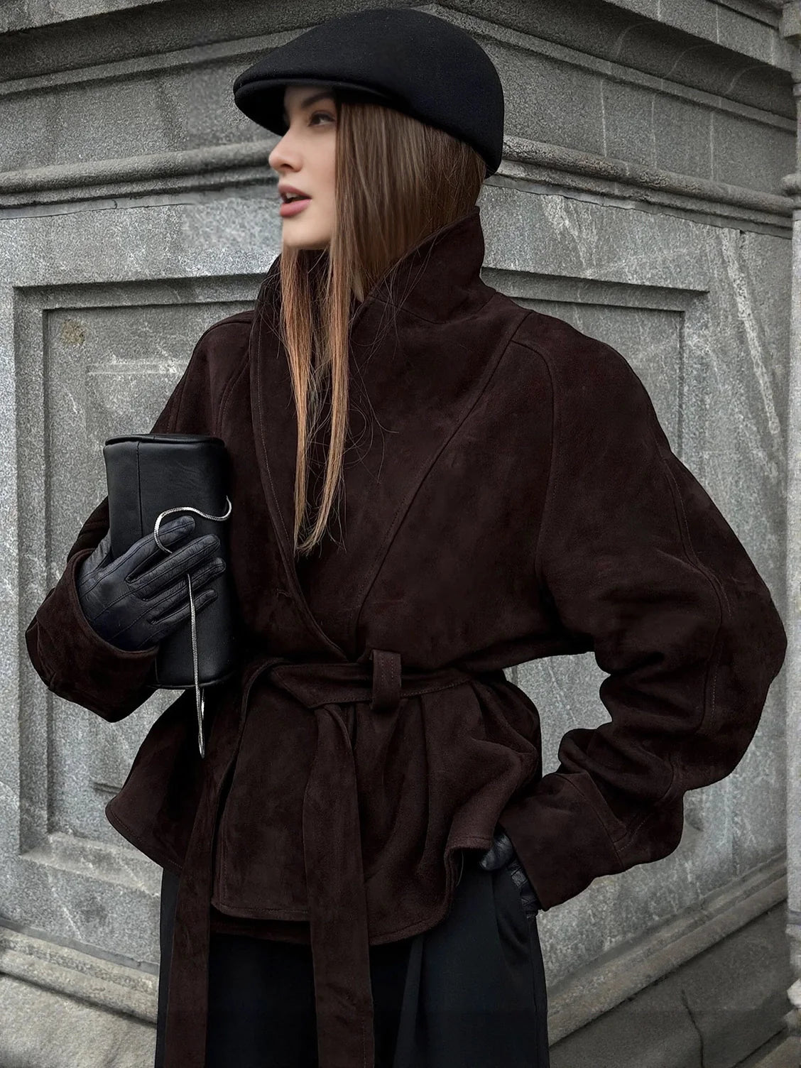 Woman in a brown coat and black hat standing against a gray wall. #Color_Brown