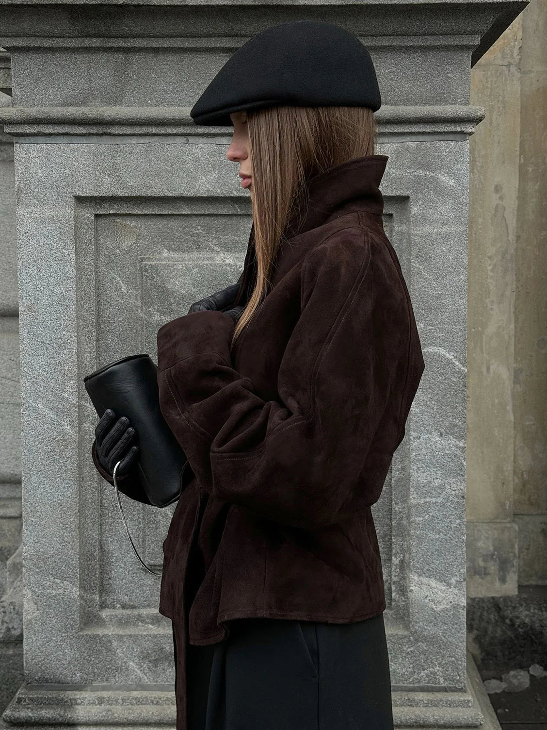 Person wearing a brown coat and black beret holding a black bag against a stone wall. #Color_Brown